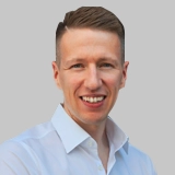 Porträt eines lächelnden Mannes mit kurzen Haaren und weißem Hemd vor grauem Hintergrund.